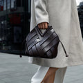 Woman holding a black woven leather handbag on city street, wearing a light coat