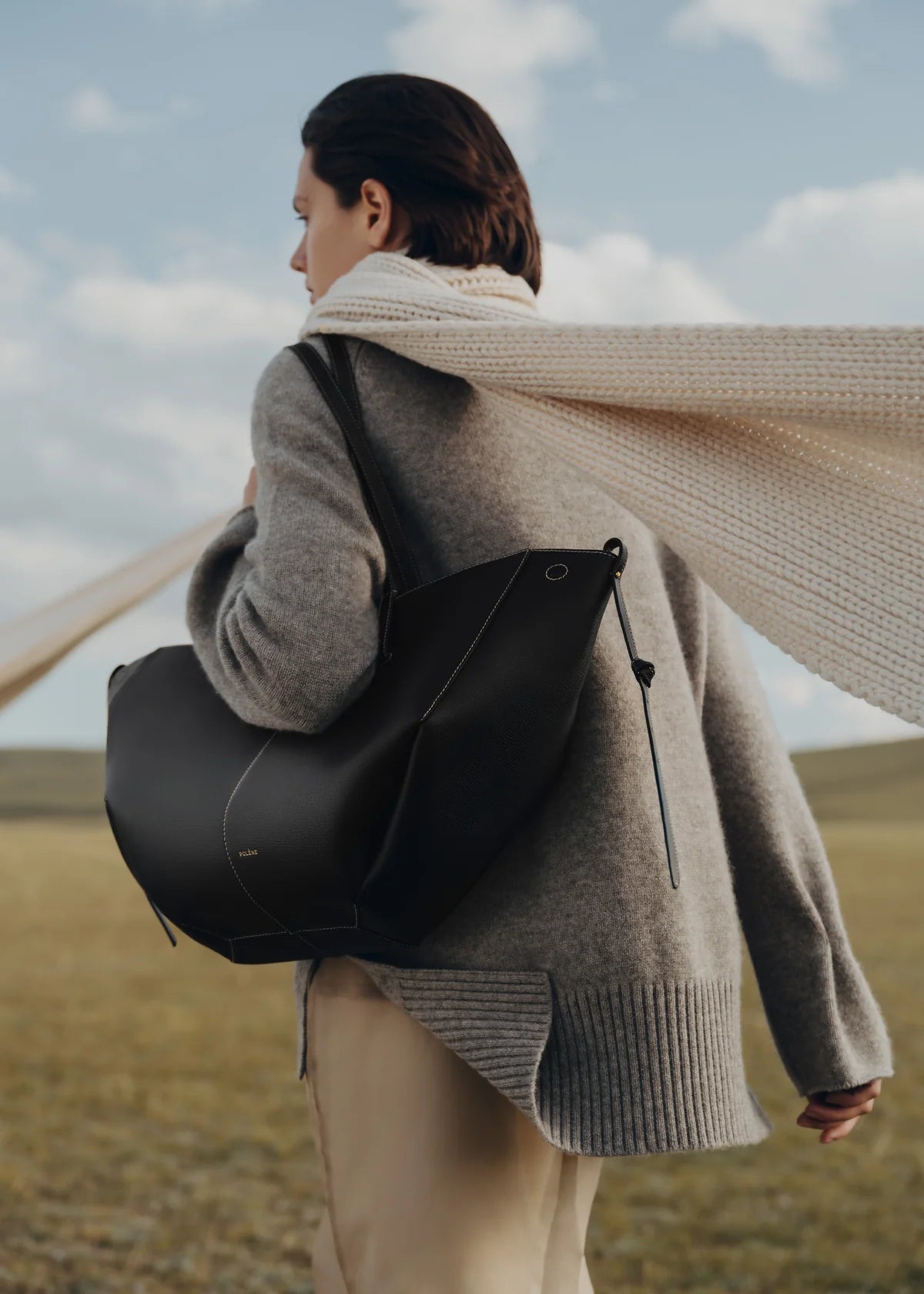 Person wearing a gray sweater and beige scarf with a black backpack in an open field.