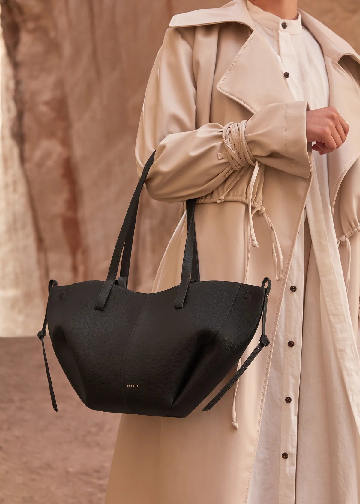 Person wearing a beige coat holding a black handbag against a neutral background